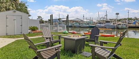 A grassy outdoor area with Adirondack chairs, a fire pit, and a marina with boats in the background.