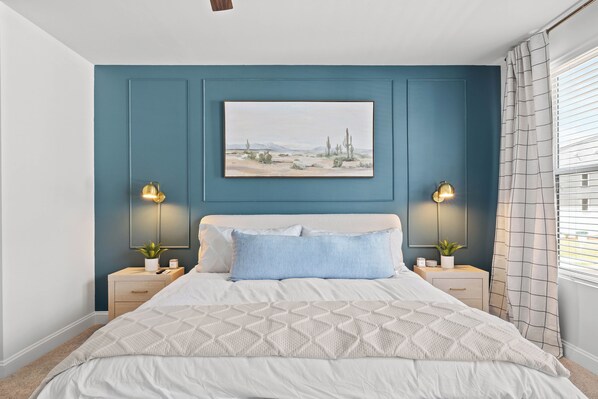 A bedroom with a bed, two bedside tables, a nightstand, a wall-mounted picture, and a window with curtains.