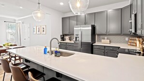 Une cuisine moderne dotée d’un grand îlot central, d’appareils électroménagers en acier inoxydable et de luminaires suspendus.