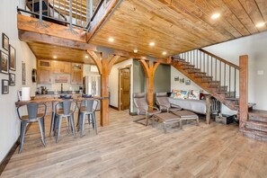 Une cuisine spacieuse avec des poutres en bois, un coin repas avec des tabourets de bar, un escalier et un lit agrémenté d’une tête de lit colorée.