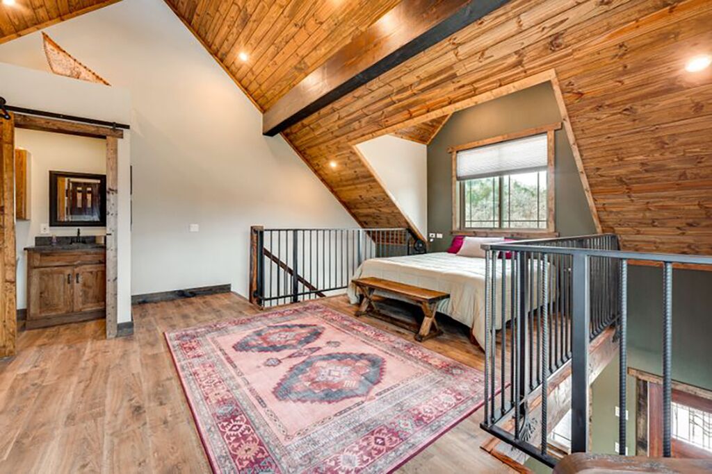 A loft bedroom with a sloped ceiling, a large window, a bed, a bench, and a patterned rug.