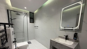 Een moderne badkamer met een douchecabine van glas, een toilet, een wastafel met een marmeren aanrechtblad en een grote spiegel met verlichte omlijsting.