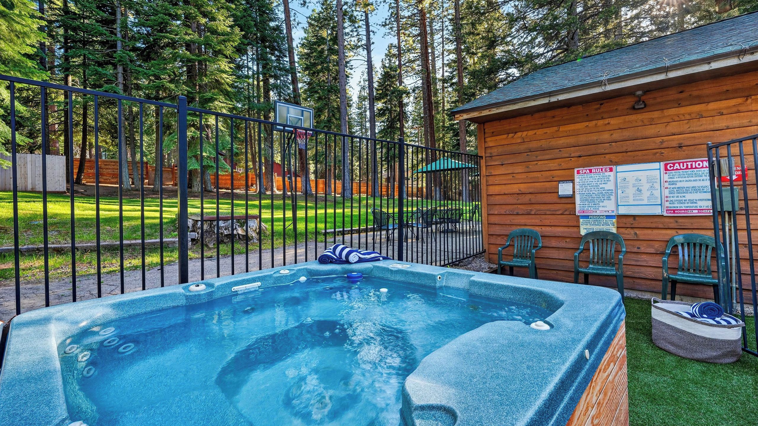 A hot tub surrounded by a wooden cabin, a fenced area, and a grassy field with trees in the background.