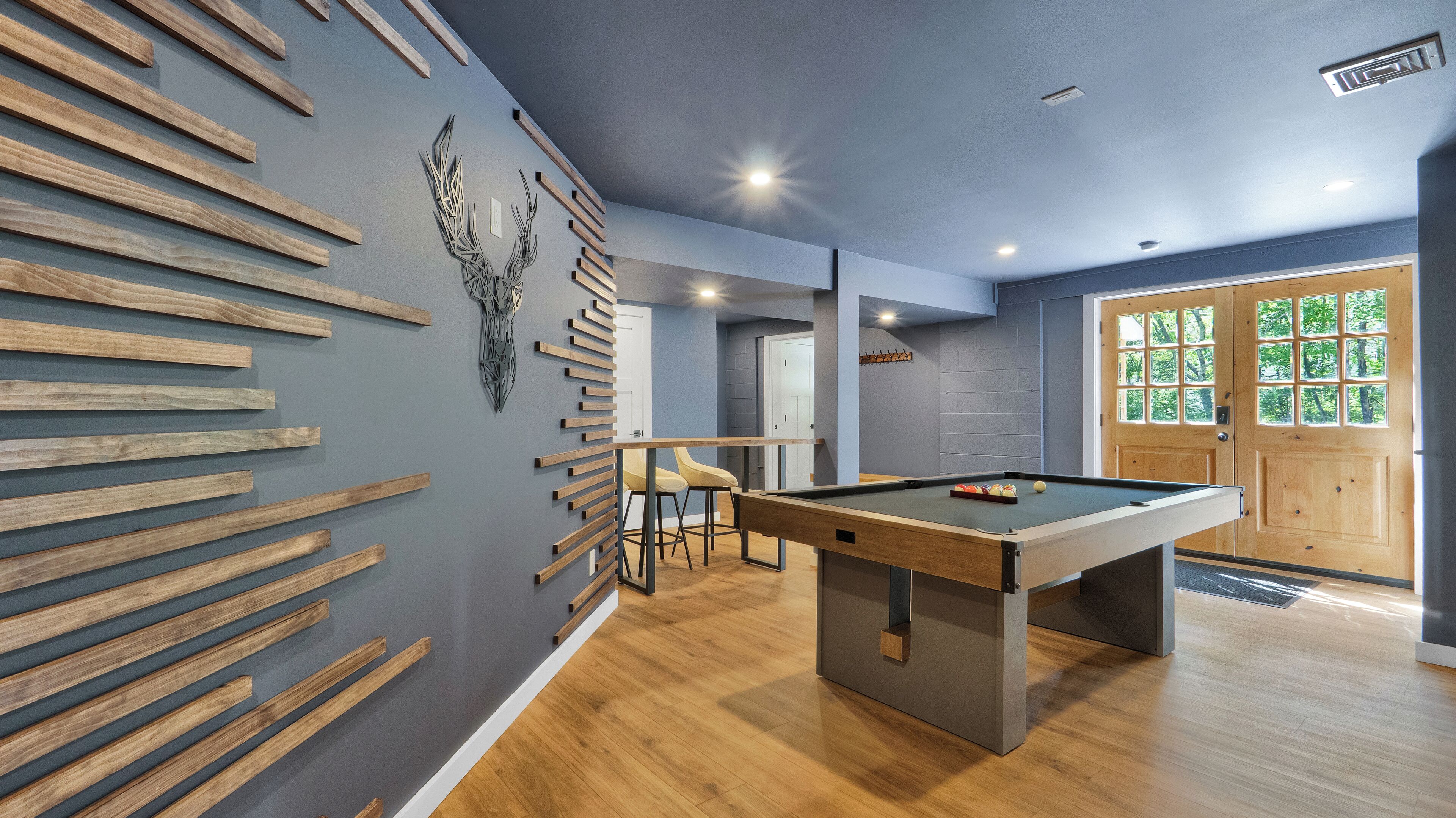 Une table de billard dans une pièce dont les murs sont recouverts de lambris en bois, avec une porte dotée d’une fenêtre et un coin bar.