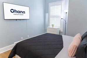 Quarto com cama, televisão na parede, mesa de cabeceira e janela com persianas.