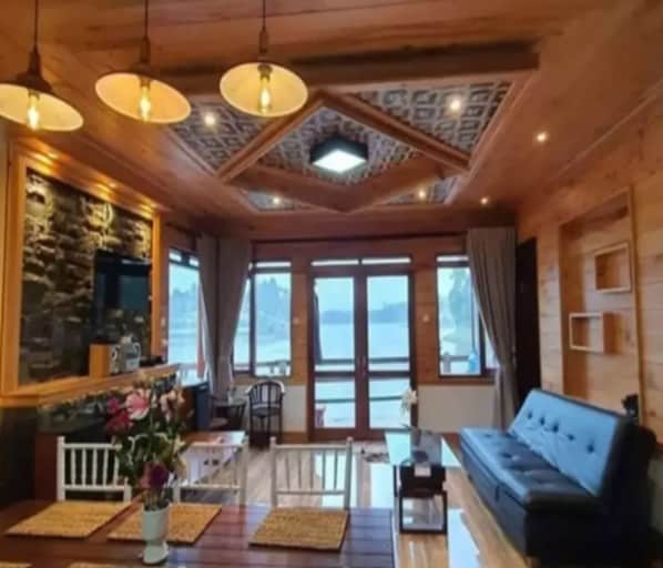 A cozy room with a wooden ceiling, a sofa, a dining table, and large windows offering a view of the outdoors. - Cozzy Villa Komando Pangalengan (Bandung)