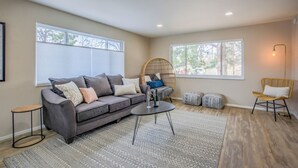 Ein Wohnzimmer mit grauem Sofa, rundem Couchtisch, Korbsessel und einem großen Fenster mit Jalousien.