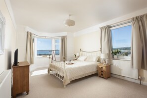 Ein Schlafzimmer mit Bett, Nachttischen, einem Schminktisch, einer Heizung, einem Fenster mit Vorhängen und Meerblick.