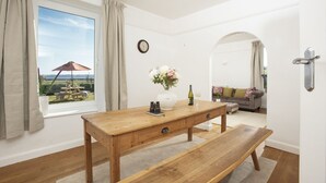 A room with a wooden table, a bench, a vase with flowers, a wine bottle, and a view of an outdoor patio. - Folneth - Parking, Garden, Beachfront (Marazion)