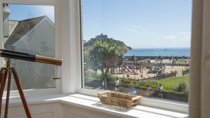 A window with a telescope, a wicker basket, and a view of a beach with playgrounds and a castle in the distance. - Folneth - Parking, Garden, Beachfront (Marazion)