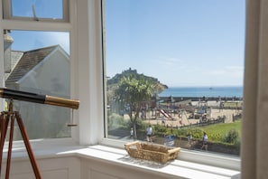 Ein Fenster mit einem Teleskop, einem Korbwesen und Blick auf einen Strand mit Spielplätzen und einer Burg in der Ferne.