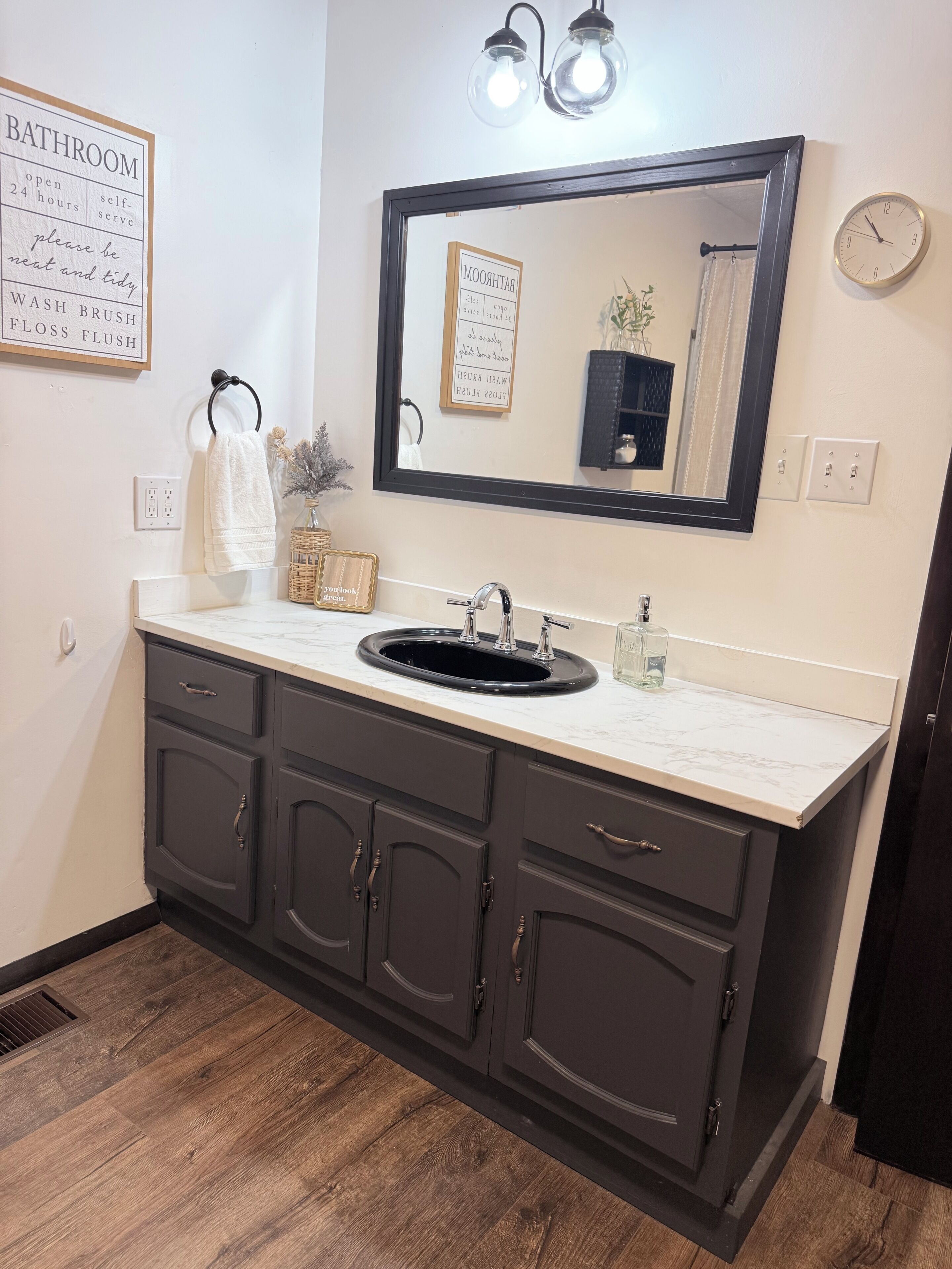 A bathroom with a sink, mirror, and cabinets. There are signs on the wall and a clock.