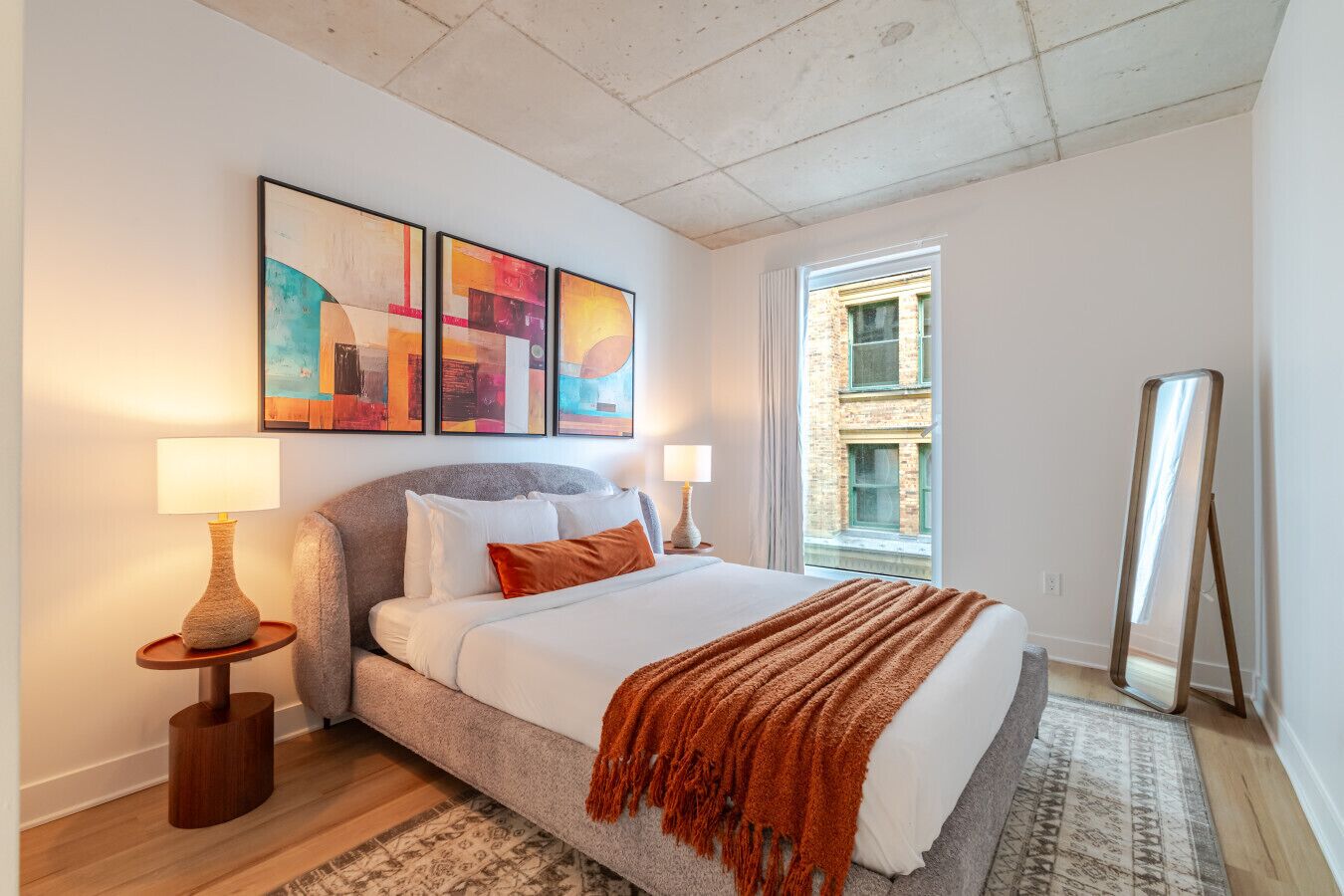 A bedroom with a bed, two bedside lamps, a rug, a window, and artwork on the wall.