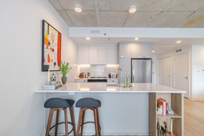 A modern kitchen with white cabinetry, a stainless steel refrigerator, and a central island with bar stools. - Old Port 2BR | Pool Gym Parking (Montreal)