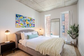 Une chambre à coucher avec un lit, des lampes de chevet, un tableau abstrait aux couleurs vives, une plante en pot et une vue sur un mur de briques à travers une fenêtre.