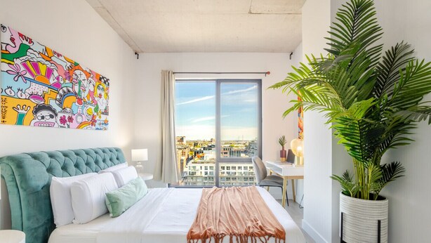 Una habitación de hotel moderna con una cama grande, vistas a la ciudad, un balcón y un mural colorido en la pared.