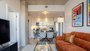 Una sala moderna con un sofá, una mesa de centro y un televisor. Se observa una cocina al fondo con un comedor.