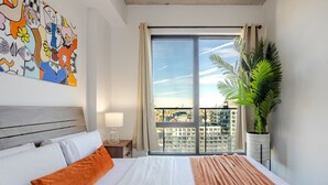 Habitación de hotel con una cama grande, una mesita de noche, una lámpara, un ventanal con vistas a edificios y una planta en maceta.