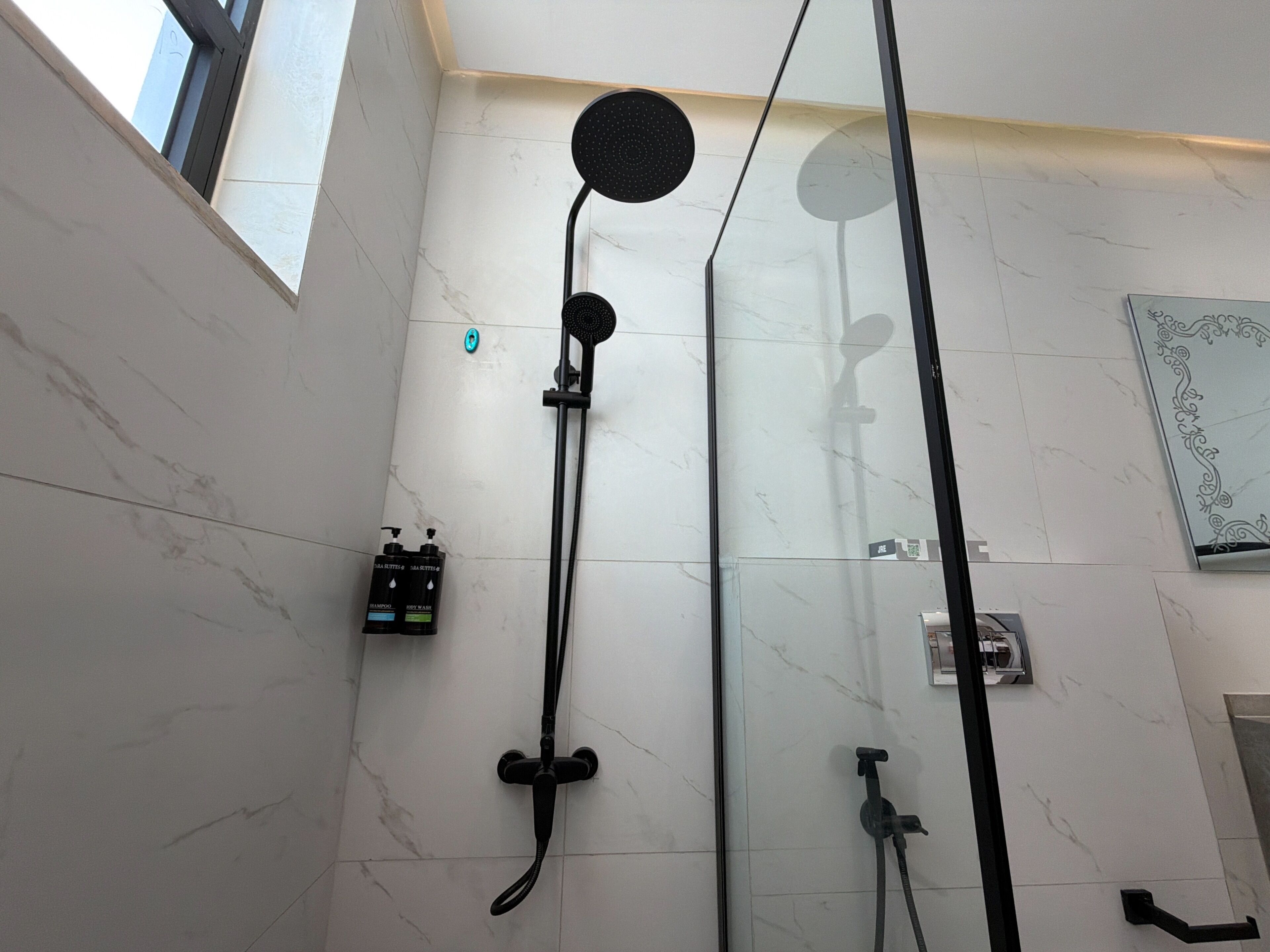 A modern bathroom with a glass-enclosed shower, a handheld showerhead, a wall-mounted faucet, and a small bottle of shampoo.
