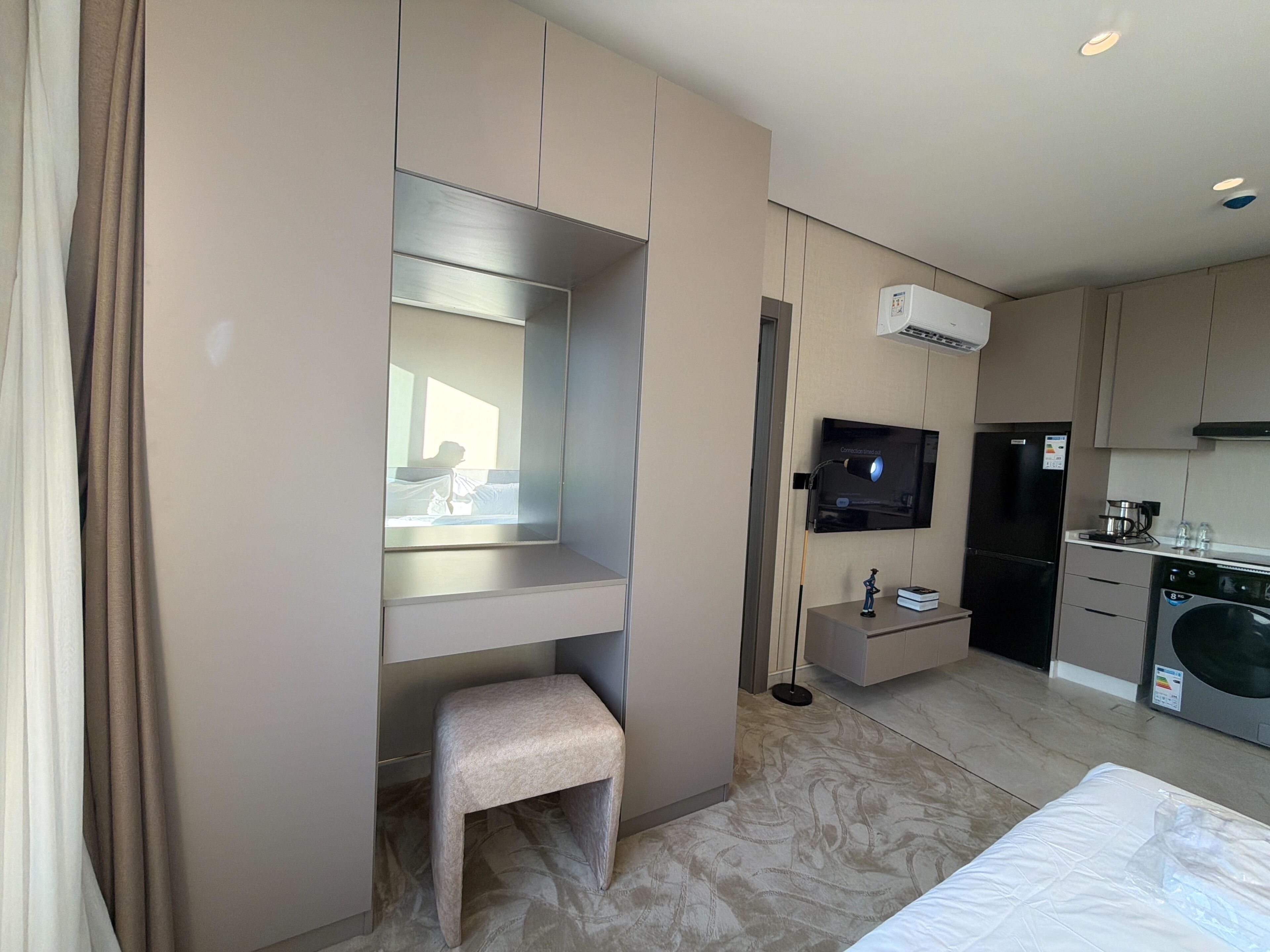 Ein Hotelzimmer mit Bett, einem kleinen Hocker, einem Fernseher, einer Waschmaschine und einer Küchenzeile.
