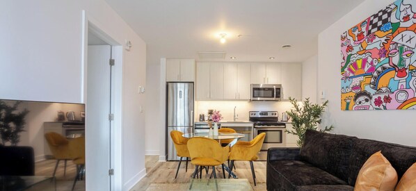 Una cocina moderna con armarios blancos, un refrigerador de acero inoxidable y un microondas. Hay una zona de comedor con una mesa de vidrio y sillas amarillas. Un sofá de color oscuro está en la sala de estar, y un colorido cuadro abstracto adorna la pared.