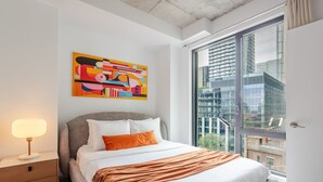 Una habitación de hotel moderna con una cama grande, una mesita de noche, una lámpara, un cuadro abstracto de colores y vistas de la ciudad a través de ventanas amplias.