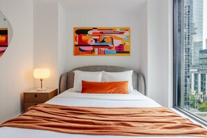 Una habitación de hotel moderna con una cama, una mesita de noche con lámpara, un cuadro abstracto de colores y vistas a la ciudad.