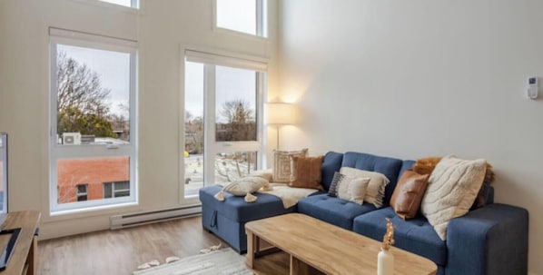 A living room with a blue sofa, a wooden coffee table, and a large window with a view of buildings and trees. - MTL Trendy Hideaway in Rosemont (Montreal)