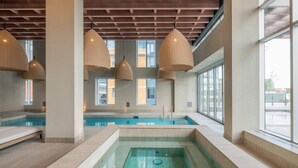 Piscina interior con vistas al exterior a través de amplios ventanales, vigas de madera en el techo e iluminación colgante.