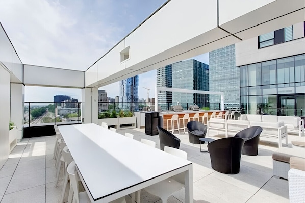 A rooftop terrace with a long white table, several chairs, and a bar area. - Charming 2BR Apt in DT MTL (Montréal)