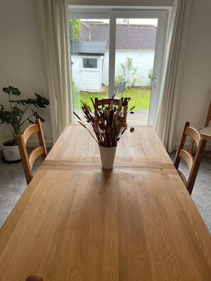 A wooden dining table with a vase of dried plants in the center, flanked by chairs, in a room with a view through a glass door to a house and garden. - Family Beach Home in Highcliffe, Dorset / New Forest with 10 Min walk to beaches (Christchurch)