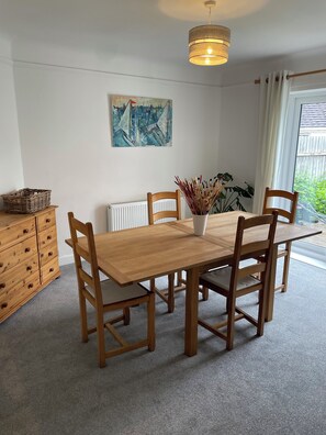 Uma área de refeições com mesa e cadeiras de madeira, luminária pendente, radiador, pintura na parede e janela com cortinas.