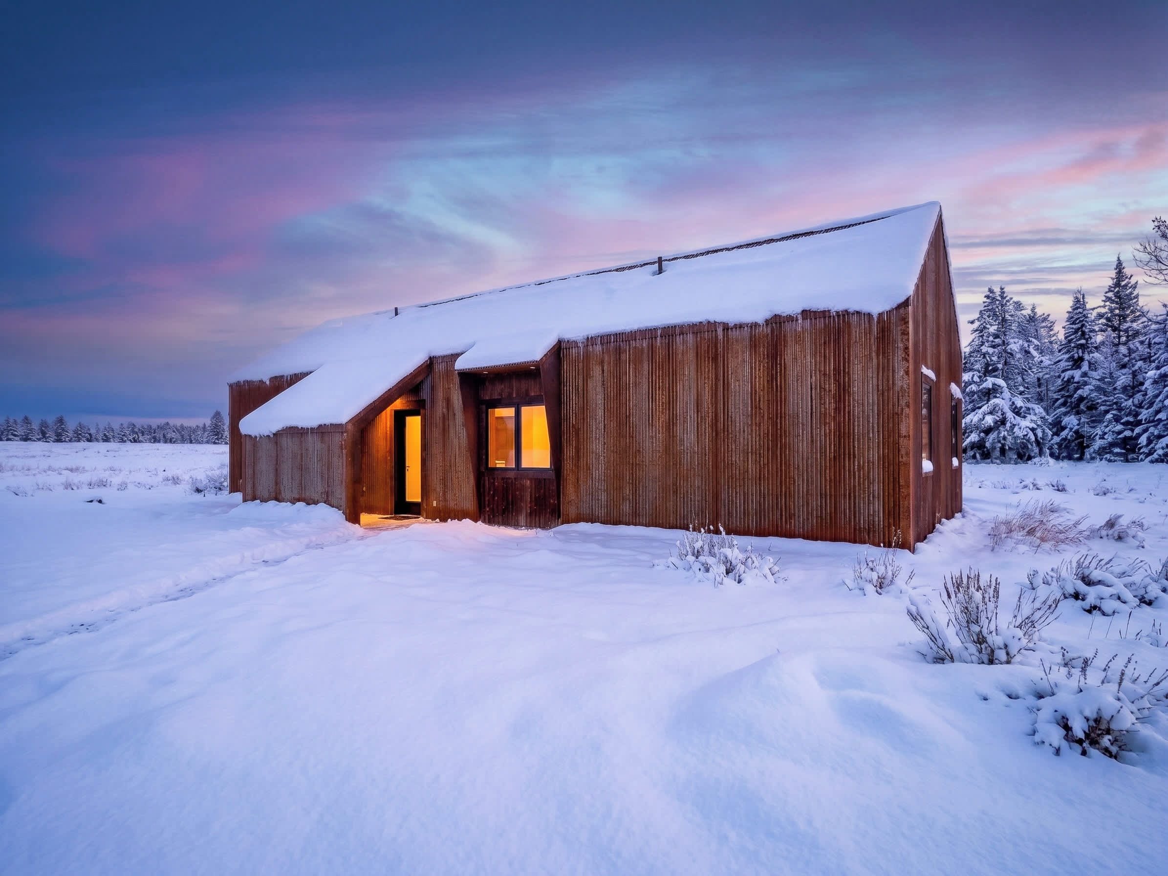 A rustic wooden cabin with a snow-covered roof, a single illuminated window, and a snowy landscape.