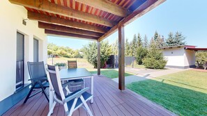 Een terras buiten met een tafel en stoelen, een houten pergola en uitzicht op een achtertuin met gazon en bomen.