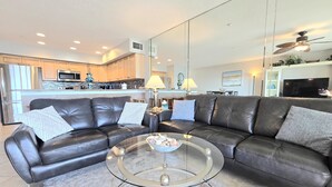 Ein modernes Wohnzimmer mit einem ledernen Ecksofa, einem Glastisch und einer Küche im Hintergrund.