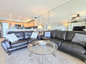 Ein modernes Wohnzimmer mit einem ledernen Ecksofa, einem Glastisch und einer Küche im Hintergrund.