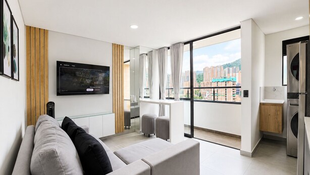 A modern apartment with a sofa, a TV, a balcony with a city view, and a kitchenette. - Apartaestudio Luxury (Sabaneta)