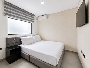 Una habitación de hotel moderna con cama, mesita de noche, lámpara, equipo de aire acondicionado y un televisor montado en la pared.