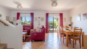 Ein geräumiges Wohnzimmer mit einem roten Sofa, einem Esstisch mit Stühlen, einer Treppe und Blick ins Freie durch große Fenster.