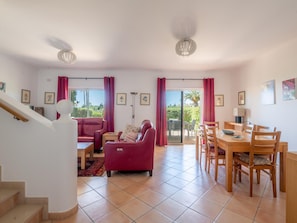 Ein geräumiges Wohnzimmer mit einem roten Sofa, einem Esstisch mit Stühlen, einer Treppe und Blick ins Freie durch große Fenster.
