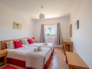 Ein Doppelbett mit roter und weißer Bettwäsche, ein Nachttisch, ein Schreibtisch, ein Stuhl, ein Fenster mit Vorhängen und eine Hängelampe.