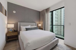 A modern hotel room with a large bed, two bedside tables with lamps, a large window with a view of buildings, and a grey textured wall.