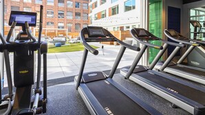 Een sportschool met loopbanden en een hometrainer, grote ramen en uitzicht op gebouwen.