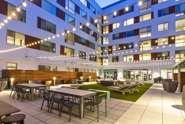 A modern hotel courtyard with outdoor seating, a grassy area, and string lights. - Modern Apartment | Gym + Courtyard (Cambridge, MA)
