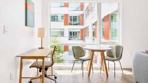 Ein modernes Zimmer mit Schreibtisch, zwei Stühlen, einer Lampe und einem großen Fenster mit Blick auf ein Gebäude.