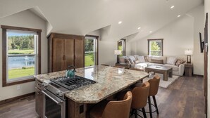 A modern kitchen with a large island, a dining area with bar stools, a living room with a sofa and a TV, and a view of a lake. - Strawberry Moon in Whitefish with Hot Tub (Whitefish)