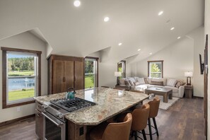 A modern kitchen with a large island, a dining area with bar stools, a living room with a sofa and a TV, and a view of a lake. - Strawberry Moon in Whitefish with Hot Tub (Whitefish)