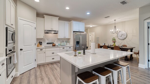 Una cocina moderna con armarios blancos, una isla central con taburetes, un comedor con candelabro y un reloj en la pared.