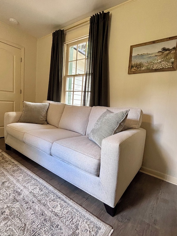 Un sofá color beige con almohadones, un tapete estampado, una ventana con cortinas y un cuadro en la pared.