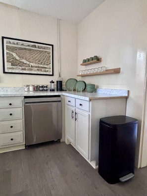 Una cocina con armarios blancos, lavavajillas, cubo de basura y un cuadro en la pared.